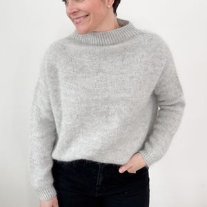 May include: A light gray sweater with a high neckline. The sweater has long sleeves and a slightly oversized fit. The person is wearing black pants. The background is a plain white wall.