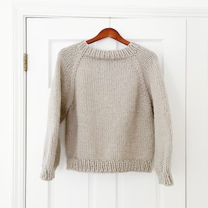 May include: A light beige knit sweater with a crew neck and long sleeves hangs on a wooden hanger. The sweater has a ribbed hem and cuffs. The background is a white door.
