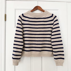 Op de afbeelding: Een gestreepte trui met een crèmekleurige achtergrond en donkerblauwe strepen. De trui heeft een ronde hals en lange mouwen.