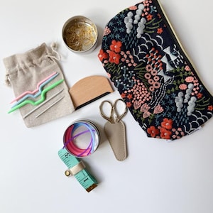 May include: A collection of sewing and knitting supplies including a dark blue floral embroidered pouch, a beige linen drawstring bag, gold-tone scissors in a tan sheath, a wooden tool, metal tins with stitch markers, and a 3-inch measuring tape.