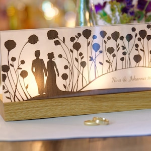 Puede incluir: Un portavelas de vidrio blanco con una silueta de una pareja de pie en un campo de flores. El texto "Nina & Johannes 16.7.2016" está impreso en la parte inferior del portavelas.