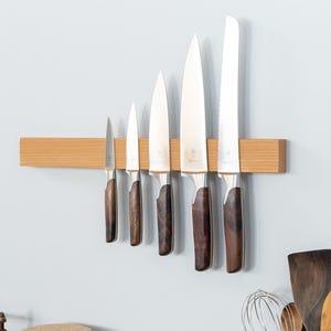 Könnte beinhalten: Eine rechteckige, horizontal montierte magnetische Messerleiste aus hellem Holz, an der fünf Küchenmesser aus Edelstahl mit dunkelbraunen Holzgriffen in unterschiedlichen Größen hängen, die von links nach rechts der Größe nach angeordnet sind.