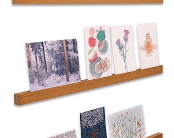 May include: Three horizontal light brown wooden wall shelves with visible layered edges, each holding a collection of rectangular greeting cards featuring various artistic prints, including floral patterns, landscapes, and botanical illustrations.
