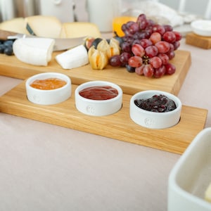 Può includere: Un vassoio di servizio in legno con tre piccole ciotole bianche riempite di diversi tipi di marmellata. Le ciotole sono disposte in fila sul vassoio. Il vassoio è in legno chiaro e ha una finitura naturale.