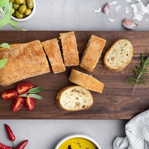 Könnte beinhalten: Ein Holzbrett mit einem in Stücke geschnittenen Brotlaib, Tomaten, Rosmarin und Knoblauchzehen.