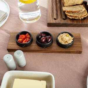 Puede incluir: Una bandeja de servir de madera con tres pequeños cuencos negros llenos de tomates cherry rojos, aceitunas negras y una crema blanca. La bandeja está sobre un mantel rosa con saleros y pimenteros, un vaso de agua y una mantequillera.