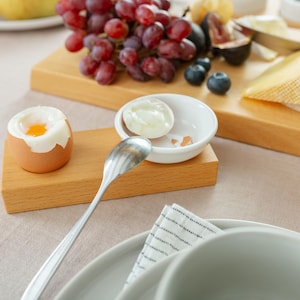 Può includere: Un primo piano di una ciotola di ceramica bianca con un uovo sodo all'interno. La ciotola è su un supporto di legno accanto a un cucchiaio d'argento. L'uovo è in un porta uova bianco.