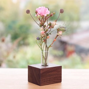 Könnte beinhalten: Eine kleine Glasvase mit rosa Rosen und anderen zarten Blumen. Die Vase steht auf einem dunkelbraunen, quadratischen Holzsockel. Der Hintergrund ist verschwommen, mit Blick auf Grün.