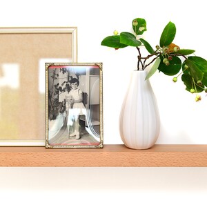 Peut inclure: Une étagère en bois avec un vase blanc rempli de feuilles vertes et de baies, un cadre photo blanc avec un passe-partout marron et un cadre photo doré avec une photo en noir et blanc d'une jeune fille.