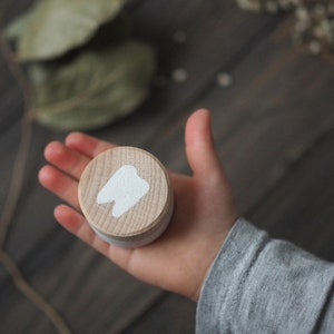 Puede incluir: Una pequeña caja de madera con un diseño de diente blanco en la tapa. La caja está siendo sostenida en una mano.