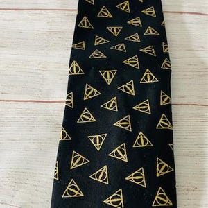 May include: Black necktie with a repeating gold Deathly Hallows symbol pattern. The tie is made of a smooth fabric and is suitable for formal or casual wear. The tie is approximately 58 inches long.