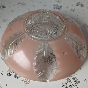 May include: A vintage, pink glass light fixture shade. The shade has a clear glass center and clear glass feather designs. The shade is round and has three small holes. The light fixture is likely from the mid-century.