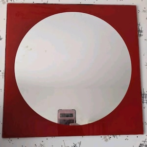 May include: Square red framed mirror with a large circular mirror in the center. The mirror reflects a phone. The frame is a solid, glossy red. The mirror is a simple, modern design.