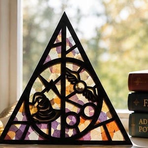 May include: A triangular black frame featuring a stained glass-style insert with multicoloured translucent pieces. The design includes a central vertical line, a circle, and silhouettes of a wizard hat, a winged ball, and round glasses.