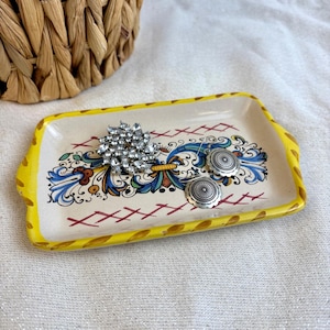 May include: A rectangular ceramic trinket tray with a yellow border and a decorative floral design in blue, green, and yellow. The tray holds a silver rhinestone brooch and two silver earrings.