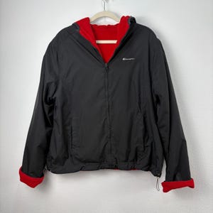 May include: Black Champion reversible jacket with a red interior. The jacket has a hood, a zip closure, and red cuffs. The Champion logo is visible on the chest. The jacket is hanging on a hanger against a white background.