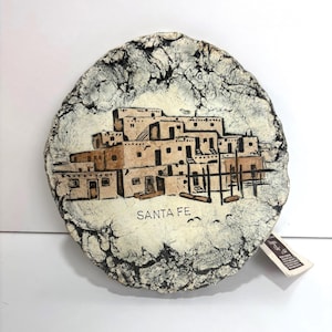 May include: A round, white ceramic plate with a black and brown painted image of a pueblo style building. The plate has a cracked, textured surface and the words "SANTA FE" are printed in black below the image.