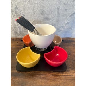 Può includere: Un pentolino per fondue in ceramica bianca con quattro ciotole per intingere più piccole in arancione, giallo, rosso e marrone. Il pentolino è su una base in legno nero e include un set di forchette per fondue.