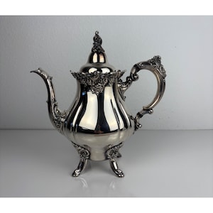 May include: A silver-colored teapot with ornate detailing. The teapot has a curved spout, a decorative handle, and sits on three small feet. The lid is topped with a decorative finial. The teapot is likely made of silver or a silver-colored metal.