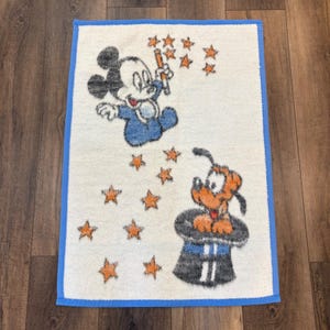 May include: A white rectangular bath mat with a blue border features a cartoon illustration of Mickey Mouse in a blue outfit, waving a wand, and Pluto in a magician's hat. Orange stars are scattered across the mat.