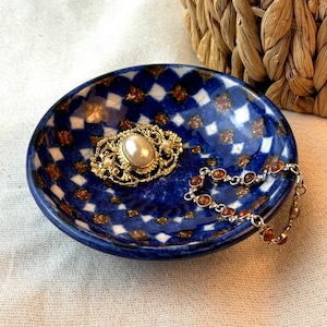 May include: A blue ceramic trinket dish with a diamond pattern, holding a gold-toned brooch with a pearl center and a silver bracelet with amber-colored stones. A woven basket is in the background.
