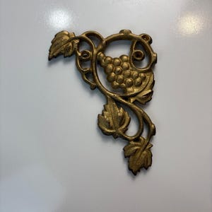 May include: Ornate gold-colored metal corner piece featuring a grape cluster and leaves. The decorative element has a detailed, vintage design, ideal for furniture or craft projects. The piece is set against a white background.