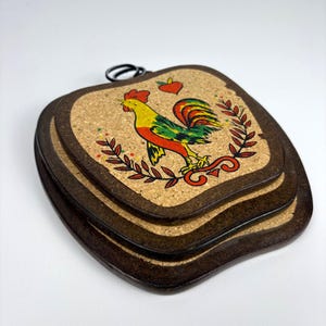 Könnte beinhalten: Ein Satz von drei dekorativen Korkuntersetzern mit einem dunkelbraunen Holzrahmen. Jeder Untersetzer zeigt eine farbenfrohe Hahn-Illustration mit roten, gelben und grünen Akzenten. Der Hahn ist von einem Lorbeerkranz und einem roten Herz umgeben.