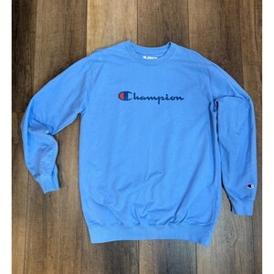 May include: A light blue Champion sweatshirt with the brand name in navy blue script across the chest. The sweatshirt has long sleeves and a crew neck. A small Champion logo is on the left sleeve.