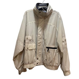 Può includere: Giacca beige con collo alto, tasche multiple e cerniera nera. La giacca ha orlo e polsini a coste. È appesa a una gruccia di metallo.