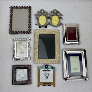 May include: A collection of decorative picture frames in various styles and sizes. Frames include silver, gold, and ornate designs, some with floral or angel motifs. One frame has the word "FAMILY" visible.