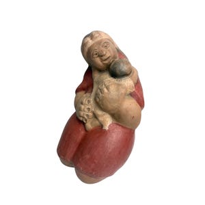 May include: A handcrafted ceramic figurine of a mother and child. The mother wears a red dress, and the baby is tan-coloured. The mother's face has a gentle expression. A unique piece of art, likely handmade.