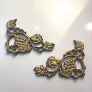 May include: Two antique brass decorative corner pieces. Each piece features a cluster of grapes, leaves, and vines. The pieces are in a gold color and are likely made of cast metal. The design is ornate and detailed.