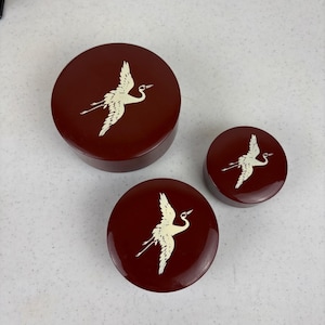 May include: Three round, burgundy-colored boxes with white crane designs. The boxes vary in size, with the largest on top and the smallest on the right. The crane is depicted in flight, with its wings spread.