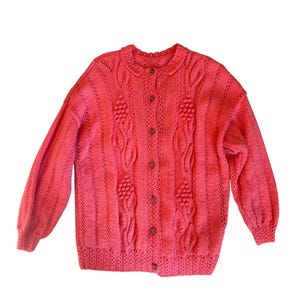 May include: A pink knitted cardigan with a button-up front and a cable knit design. The cardigan has a ribbed collar and cuffs.