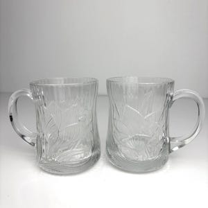 May include: Two clear glass mugs with handles. Each mug has an embossed floral design and vertical ribbed texture. The transparent mugs allow the contents to be seen. Suitable for hot or cold drinks. Each mug is approximately 10 cm tall.