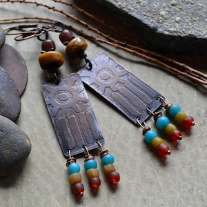 May include: Handmade copper earrings with a rectangular design and a sun-like pattern. The earrings feature colorful beads in shades of blue, yellow, and red. The earrings are suspended from copper hooks.