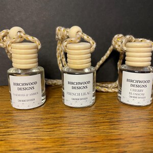 May include: Three small glass car and boat diffuser bottles with light wood caps and hanging cords. Each bottle features a white label with the text Birchwood Designs and specific scent names: Oakmoss & Amber, French Lilac, and Cherry Blossom.