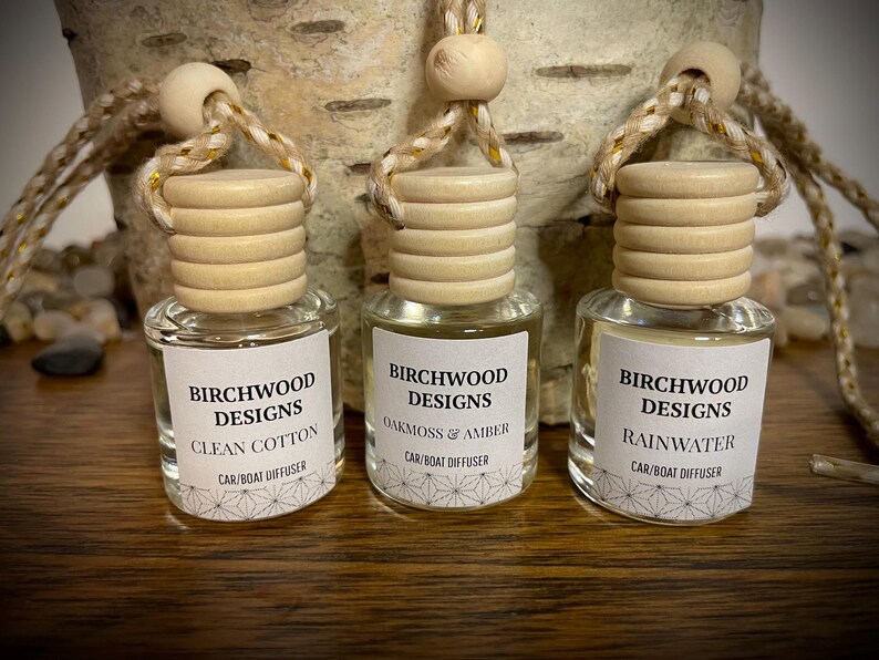 K&ouml;nnte beinhalten: Drei kleine Glas-Auto-/Bootsdiffusoren mit Holzdeckeln und Aufh&auml;ngeschn&uuml;ren. Jede Flasche hat ein Etikett mit dem Text "Birchwood Designs" und einem Duftnamen: Clean Cotton, Oakmoss & Amber und Rainwater. Der Hintergrund enth&auml;lt Birkenrinde.