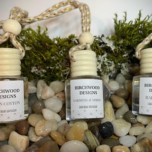 K&ouml;nnte beinhalten: Drei Birchwood Designs Auto-/Bootsdiffusoren mit Holzverschl&uuml;ssen und Etiketten. D&uuml;fte sind Clean Cotton, Oakmoss & Amber und Rainwater. Die Diffusoren h&auml;ngen an einem dekorativen Seil und sind von Kieselsteinen und Gr&uuml;n umgeben.