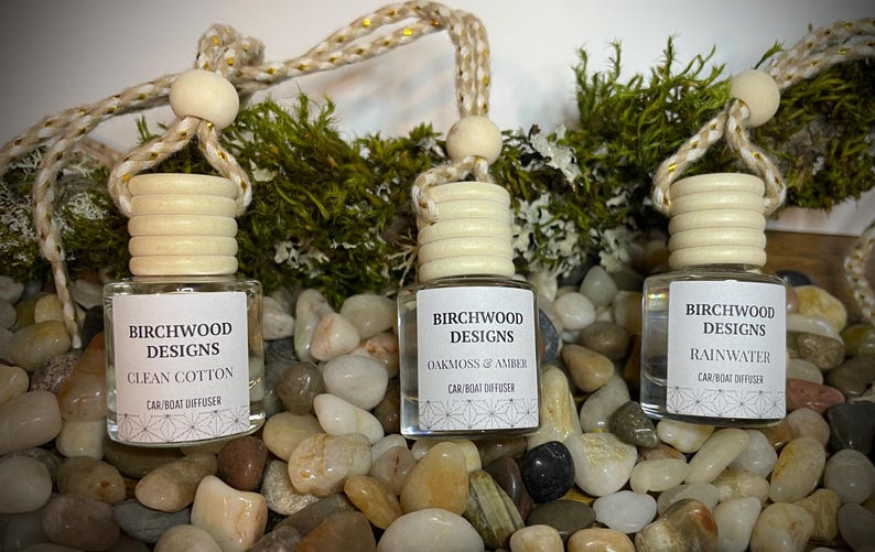 K&ouml;nnte beinhalten: Drei Birchwood Designs Auto-/Bootsdiffusoren mit Holzverschl&uuml;ssen und Etiketten. D&uuml;fte sind Clean Cotton, Oakmoss & Amber und Rainwater. Die Diffusoren h&auml;ngen an einem dekorativen Seil und sind von Kieselsteinen und Gr&uuml;n umgeben.