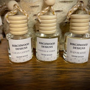 Może przedstawiać: Trzy małe szklane dyfuzory do samochodu/łodzi z drewnianymi wieczkami i sznurkami do zawieszania. Każda butelka ma etykietę z napisem "BIRCHWOOD DESIGNS" i nazwą zapachu: Clean Cotton, Oakmoss & Amber i Rainwater.