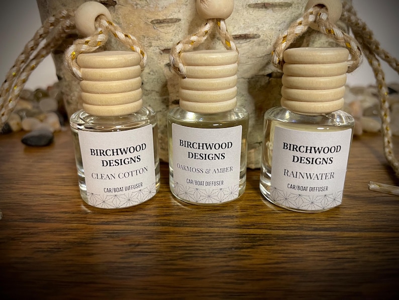 K&ouml;nnte beinhalten: Drei kleine Glas-Auto-/Bootsdiffusoren mit Holzdeckeln und Aufh&auml;ngeschn&uuml;ren. Jede Flasche hat ein Etikett mit dem Text "BIRCHWOOD DESIGNS" und einem Duftnamen: Clean Cotton, Oakmoss & Amber und Rainwater.