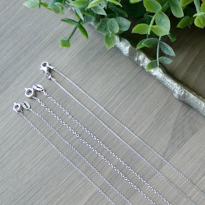 Puede incluir: Cinco cadenas de plata de diferentes longitudes dispuestas sobre una superficie de madera. Las cadenas son todas del mismo estilo y tienen un diseño simple y delicado.
