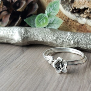 May include: A silver-colored ring with a floral design. The ring features a flower and two leaves on the band. The ring is on a wooden surface with decorative elements in the background.
