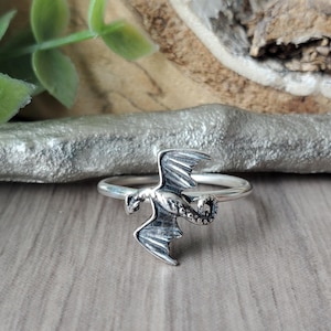 May include: A silver dragon ring with intricate detailing. The dragon has spread wings and a textured body. The ring is displayed on a wooden surface, with foliage in the background, creating a fantasy theme.