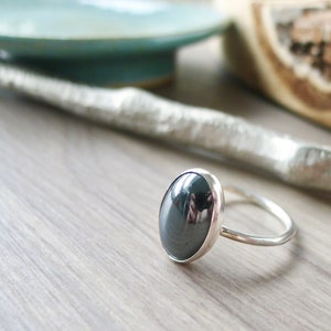 Puede incluir: Un anillo de plata con una piedra ovalada negra. El anillo está sobre una superficie de madera.