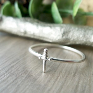 May include: A silver ring with a small sword design. The ring is thin and delicate.