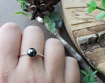 Puede incluir: Un anillo de plata con una piedra preciosa negra engastada en un engaste de bisel. El anillo está en el dedo de una persona.