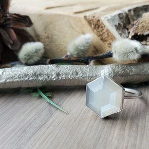 May include: A silver ring featuring a hexagonal, pale grey gemstone. The ring is set against a natural backdrop with wood and greenery. The gemstone has a faceted surface, reflecting light.