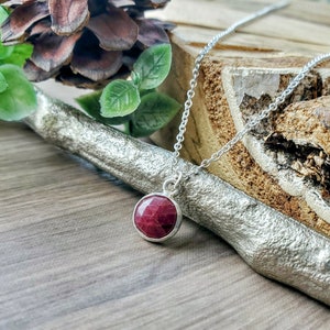 May include: A silver chain necklace with a red gemstone pendant. The pendant is round and has a faceted surface.